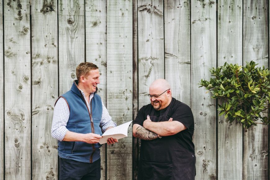 Two men stand against a wooden bar laughing and talking, the one on the left holding a menu